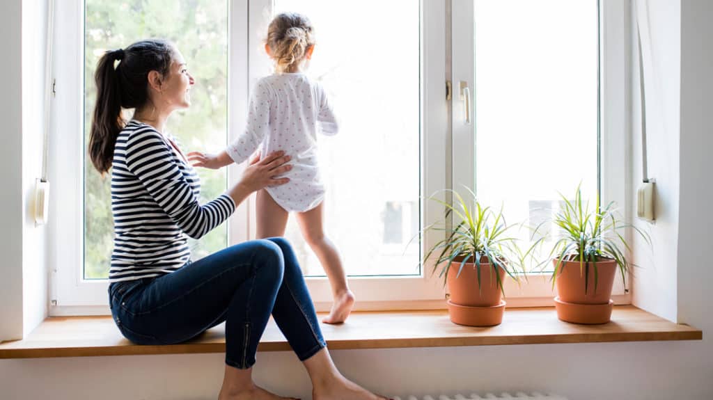 Smiling mom sitting on a window ledge, steadying her young daughter who’s standing, looking out the window