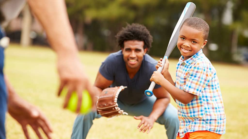 Smiling boy with a baseball bat about to swing at a pitch while his dad is playing catcher in the background
