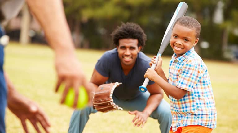 Smiling boy with a baseball bat about to swing at a pitch while his dad is playing catcher in the background