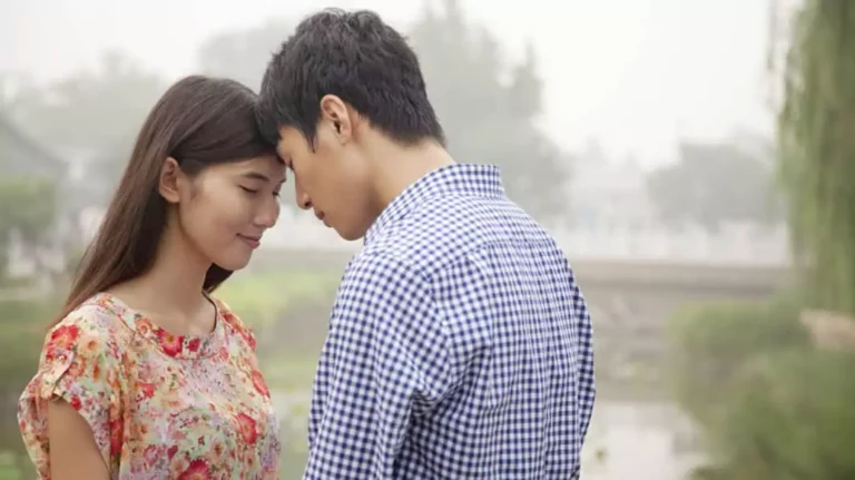 A couple stands closely with their foreheads touching in an intimate moment, symbolizing a connection that might raise considerations around topics like premarital sex. The pair appears deeply emotional and close, set against a serene, natural backdrop.