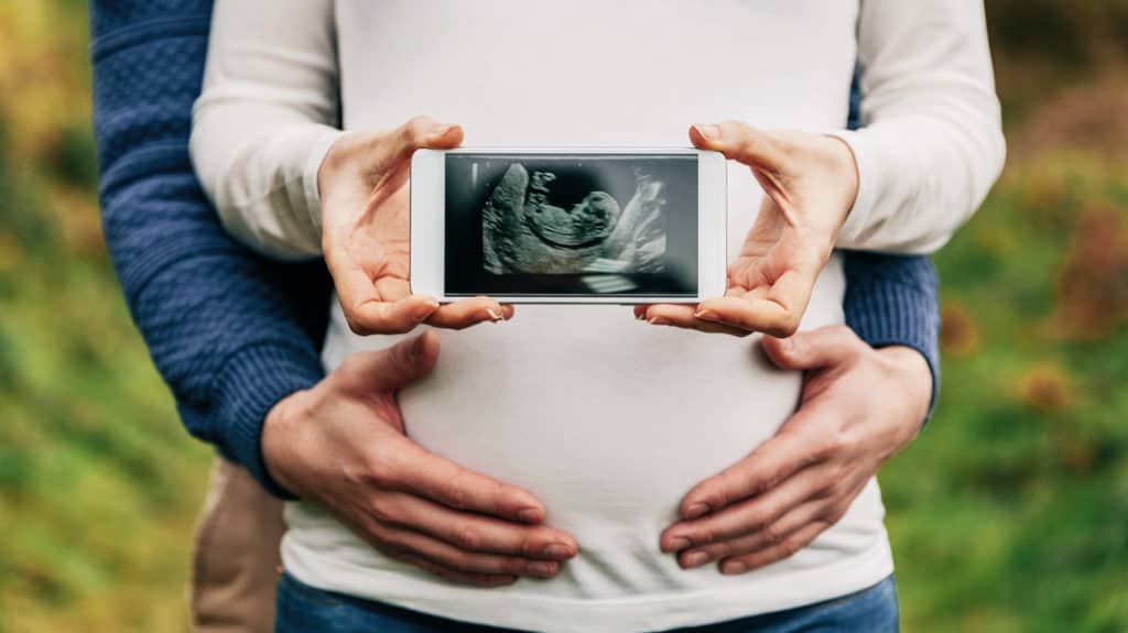 A man holds the belly of his pregnant wife. She holds a photo of a viable ultrasound.