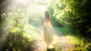 Shown from behind, a woman in a dress walking down a nature path with bright sunshine making everything appear to glow