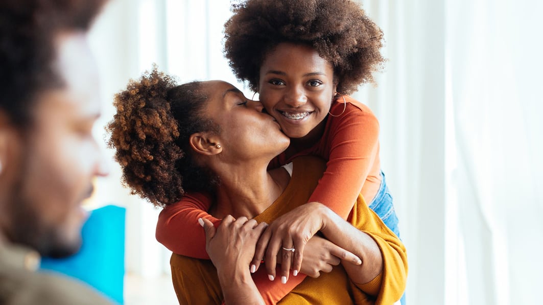 Smiling teen girl hugging her seated mom from behind, while mom kisses her cheek