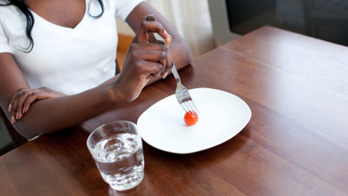 Black woman using fork to stab mini tomato on otherwise empty plate next to a glass of water, implying an eating disorder