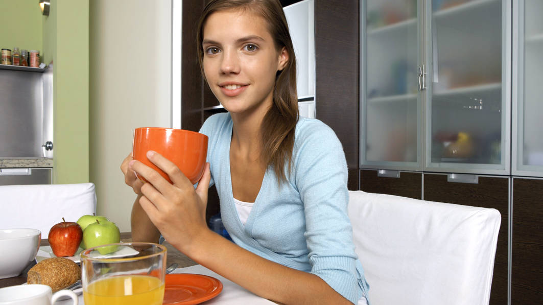 A slight smile while looking at the camera, a thin teen girl sits at a breakfast table, holding a large orange mug