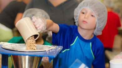 Young boy volunteering at a food bank, pouring a cup of cereal into a funnel