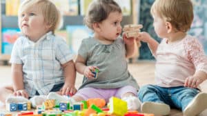 Three toddlers sit side-by-side in front of a pile of toys. One tries to take a toy airplane from the middle child.