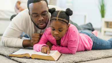 Father lying on the floor with his young daughter as she reads the Bible