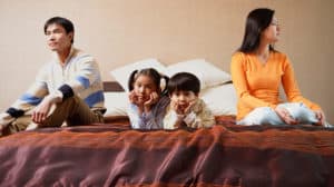Serious-looking family sitting on large bed, with the couple sitting on opposite ends and two young kids lying in the middle