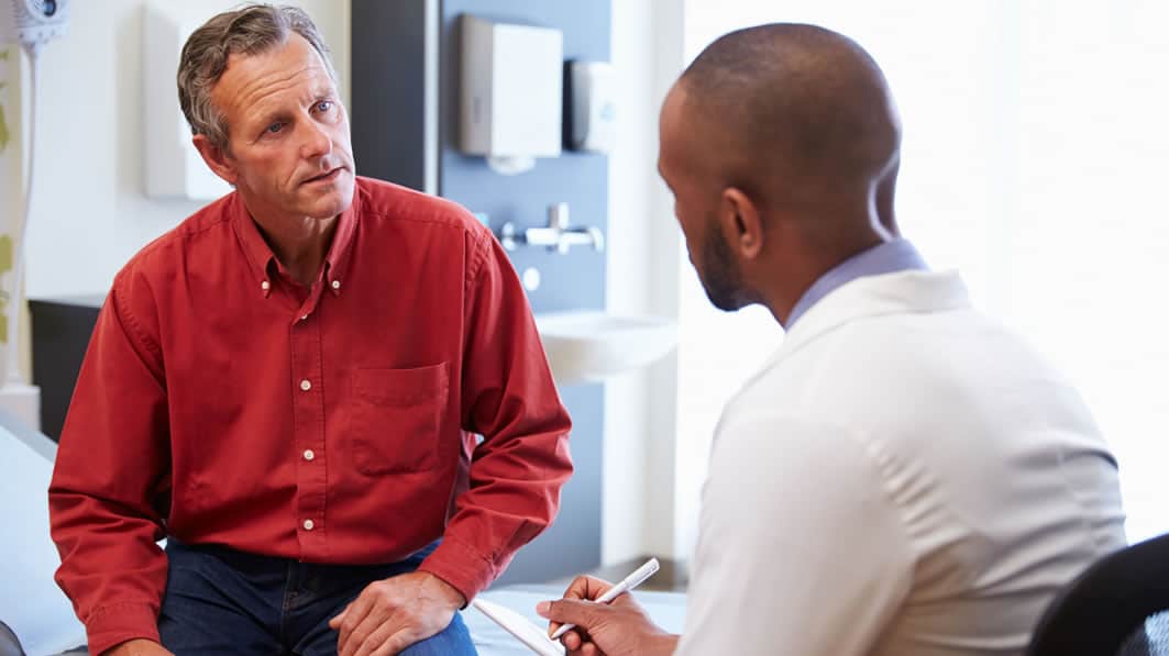 Middle-age man sitting on the edge of a bed in a hospital room, having a serious discussion with his doctor