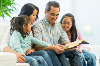 Smiling dad, mom, and two young daughters sitting on the couch studying the Bible