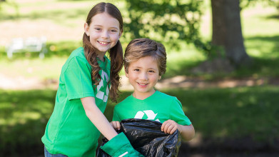 Two young kids smiling for the camera, wearing green shirts with the recycling logo and picking up trash in a park