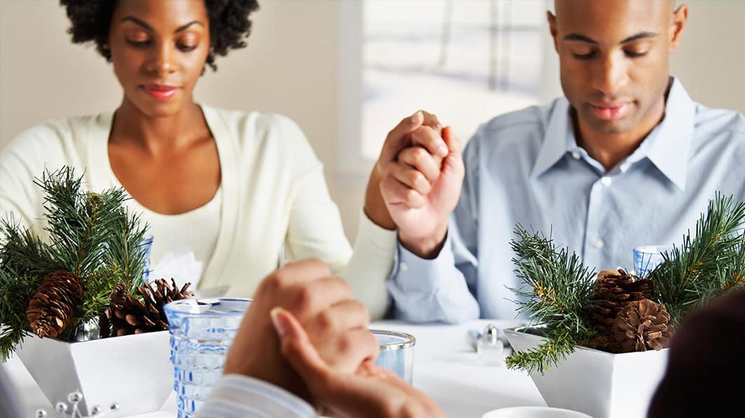 Family saying grace around the dinner table, with focus on one couple holding hands