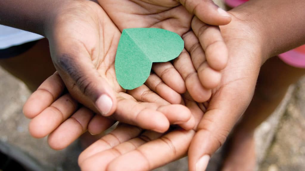 Close up of a green paper heart held in a child's hands, and a pair of older hands beneath the child's