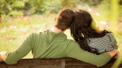 Shown from behind, a couple sitting on a bench outside. His arm is around her, their heads are leaning against each other.