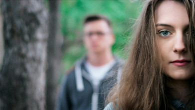Close up of young, serious-looking woman with a man standing in the blurry background, suggesting a troubled relationship