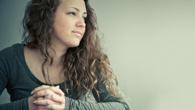 A young woman sits at a table with her hands folded atop a Bible as she looks reflectively to her left