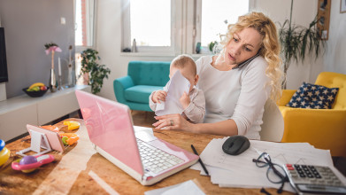 Busy, stressed-out mom holding her baby in her lap while she’s talking on the phone and working on her computer