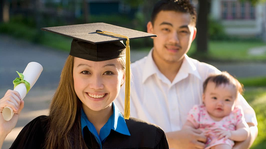 A smiling college graduate holding up her new degree, her husband cradling their baby in the background