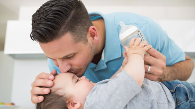 Young father affectionately resting his hand on and kissing his baby’s forehead as he gives the boy his feeding bottle