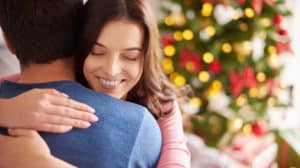 Close up of a smiling wife's face as she's hugging her husband with a Christmas tree in the background