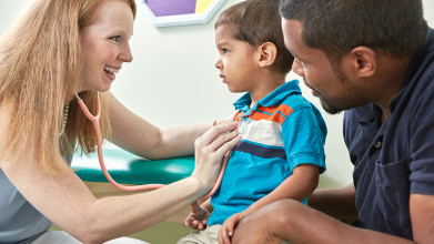 Smiling female doctor checking a young boy's heart with a stethoscope as his father looks on