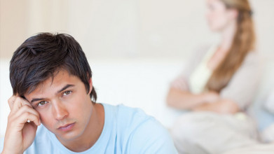 Contempative husband as frustrated wife stands in background with arms crossed