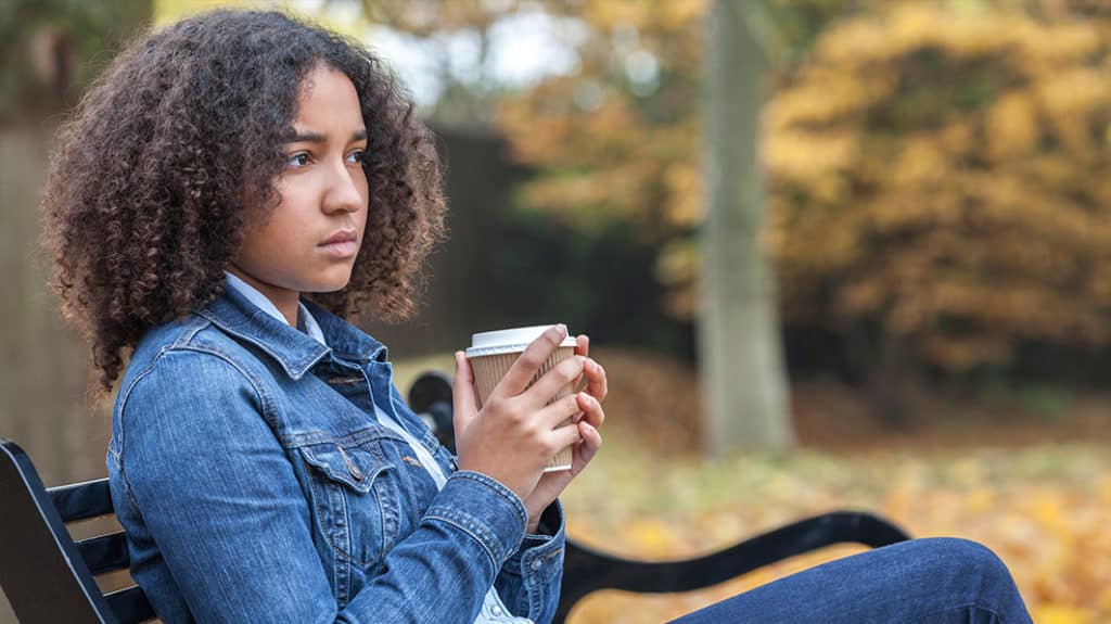 A teen thinks deeply as she sits on a park bench holding a hot drink