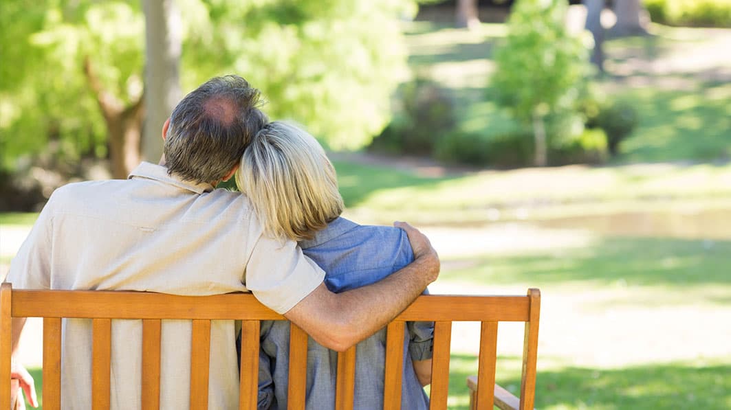 Shown from behind, a middle-age husband and wife sitting on a park bench. He has his arm around her.