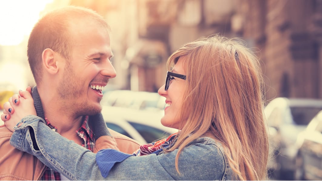 Smiling, happy couple standing on a city street. She’s got her arms around his neck as they look at each other face-to-face.