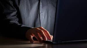 Close up of a man's hand typing on a laptop as he sits in the dark
