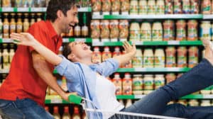 Husband and wife laugh as husband playfully pushes wife in cart inside grocery store