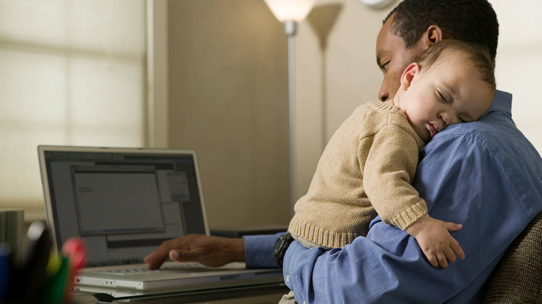 Businessman using laptop while holding infant so on his shoulder in his home office