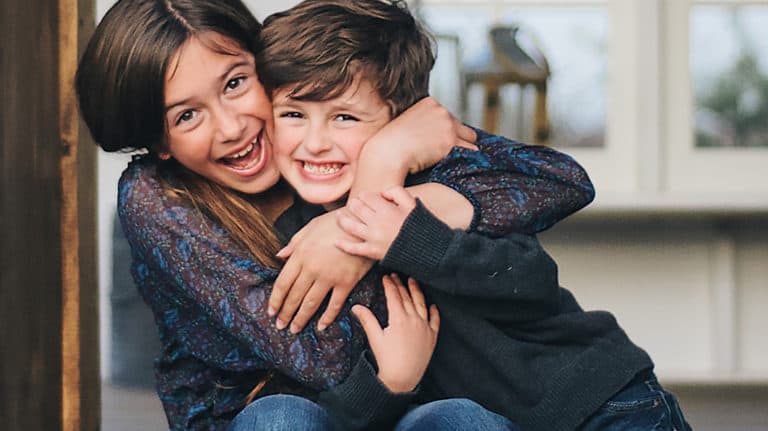 Older sister tightly hugging brother around the neck. Both are laughing.