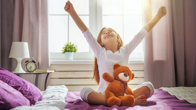 Young girl sitting on her bed with a teddy bear in her lap. She's cheerfully raising her arms in a ‘V for victory’ way