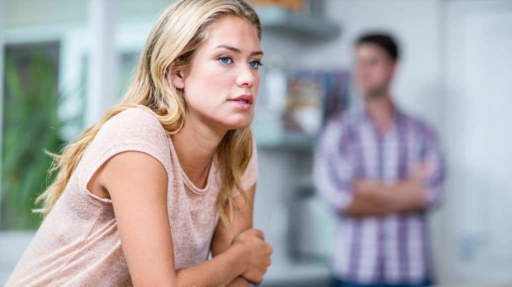 Troubled young couple. She’s in the foreground looking contemplative, he’s standing in the background with his arms crossed.