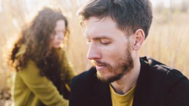 Young, somber-looking couple sitting in a field; man in foreground, woman in background