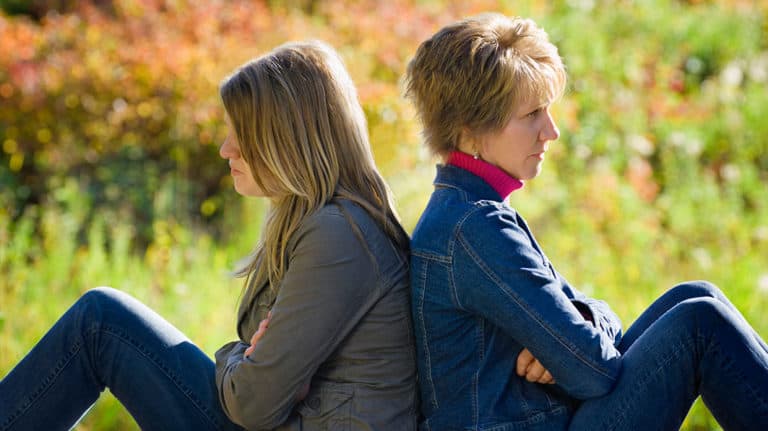 Serious-looking mom and teen daughter sitting back to back in a field