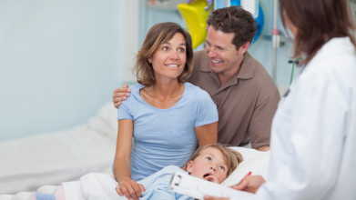 Happy parents receiving good news from a doctor about their daughter who’s lying next to them in a hospital bed