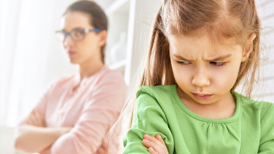 Young girl frowning and pouting, with mom in the background looking at her