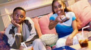 Teen brother and sister sitting on couch with snacks and watching TV; she’s got a big smile and holding up a remote
