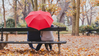 Shown from behind, a husband and wife sitting under an umbrella on a park bench on an autumn day