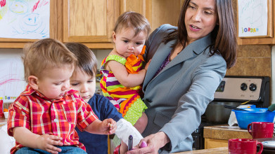 A busy mom in a business suit holding her toddler while helping her two boys with something in the kitchen