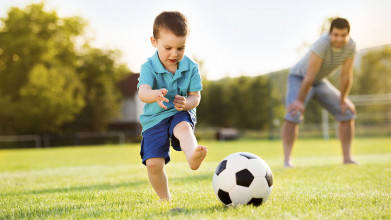 Young boy kicking a soccer ball as his smiling father watches in the background