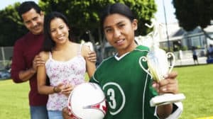 Champion girl soccer player posing for camera with her ball and trophy as her proud parents watch in the background