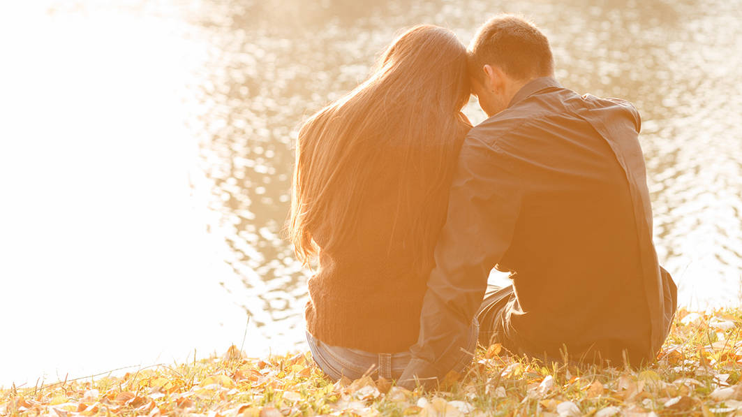 Young affectionate couple sitting by a lake, shown from behind