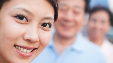 Close up of smiling Asian woman in the foreground, with her smiling parents blurred out in the background