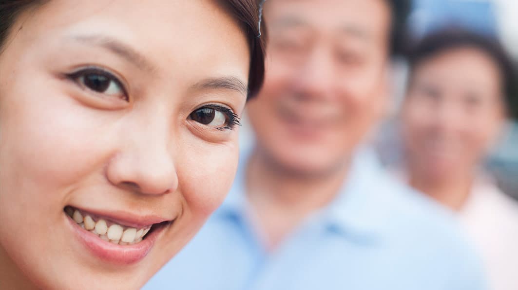Close up of smiling Asian woman in the foreground, with her smiling parents blurred out in the background