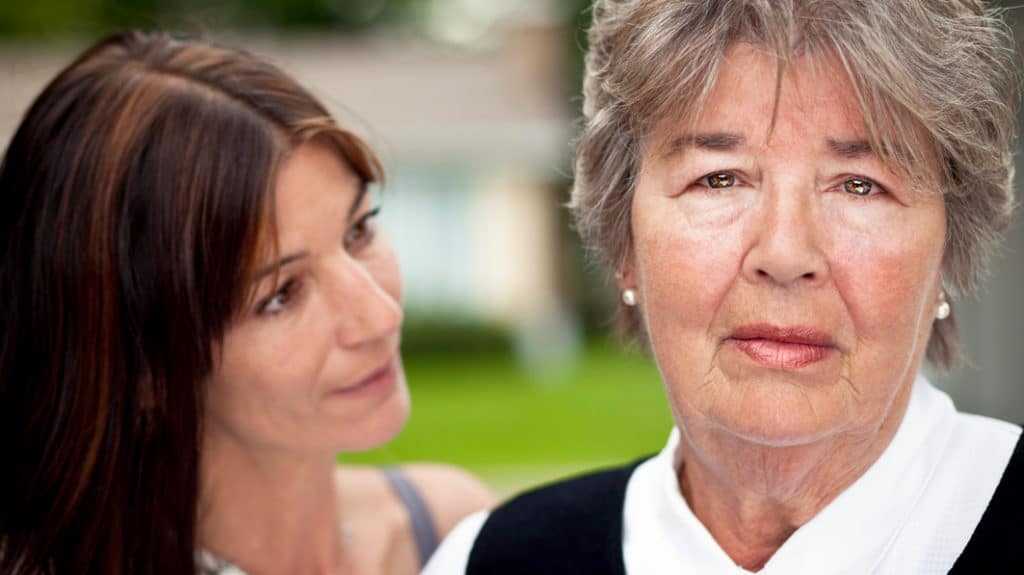 Close up of a sad mom looking into camera with her adult daughter standing beside looking at her