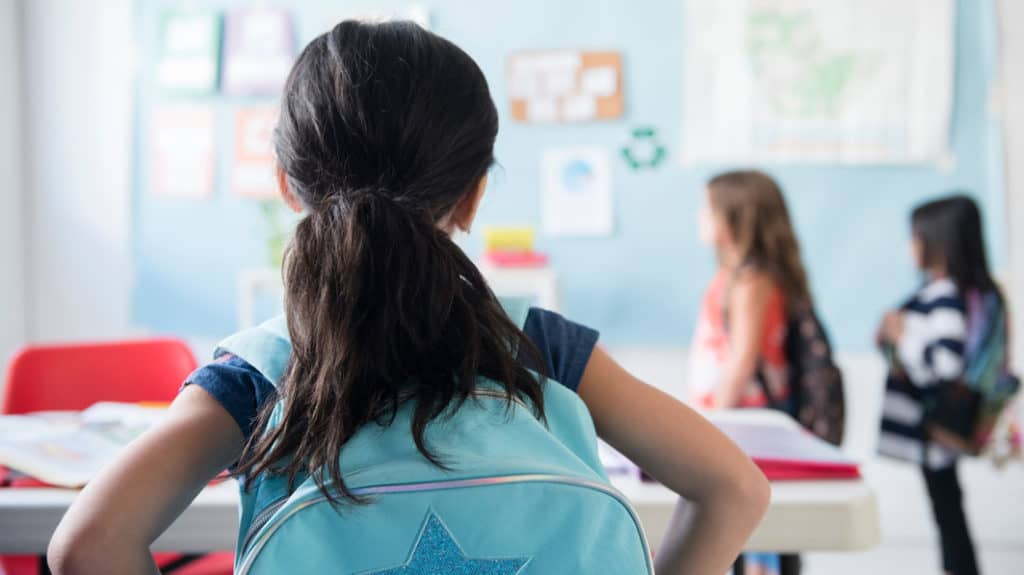 Girl in classroom wearing backpack watches classmates